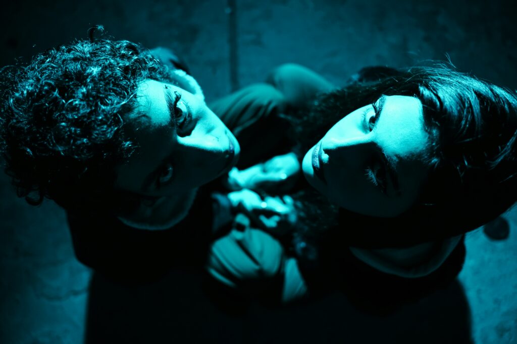 Zwei von oben fotografierte Personen, die hinauf in die Kamera schauen und in hellblaues Licht getaucht sind