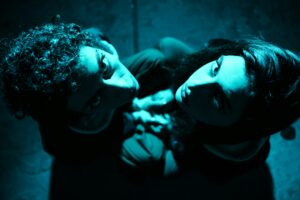 Zwei von oben fotografierte Personen, die hinauf in die Kamera schauen und in hellblaues Licht getaucht sind