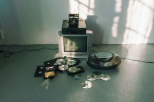 Ansammlung veralteter Medien, mittig ein Röhrenfernseher, ein CD-Player, lose verteilte CDs und eine Polaroid-Kamera