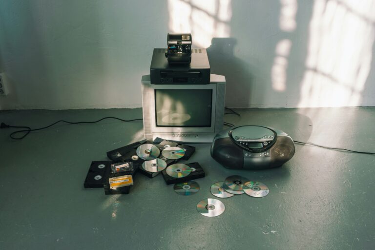 Ansammlung veralteter Medien, mittig ein Röhrenfernseher, ein CD-Player, lose verteilte CDs und eine Polaroid-Kamera