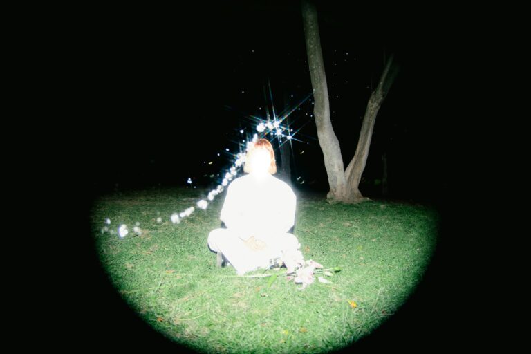 Eine überbelichtete Person, die auf einer Wiese sitzt, rechts im Bild der Stamm eines Baumes. Durch das Bild zieht sich eine geschwungene Linie aus Glitzerpunkten. Die Ränder des Bildes sind geschwärzt