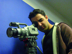 Ein junger Mann in brauner Jacke, der mit einem Auge durch eine Filmkamera schaut