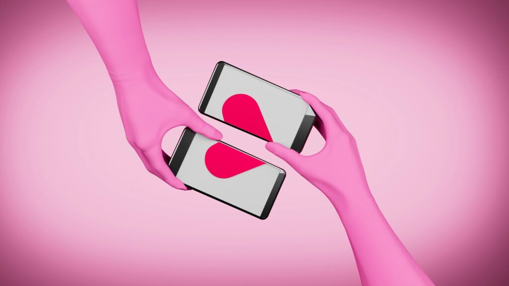 Zwei rosa Arme, die in der Mitte des Bildes zwei Handys nebeneinander halten, sodass die Bildschirme sich zu einem roten Herz ergänzen