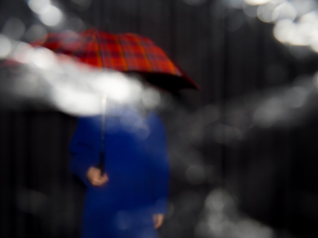 Verschwommenes Bild mt Blau gekleideterr Person mit einem roten Regenschirm überm Kopf.